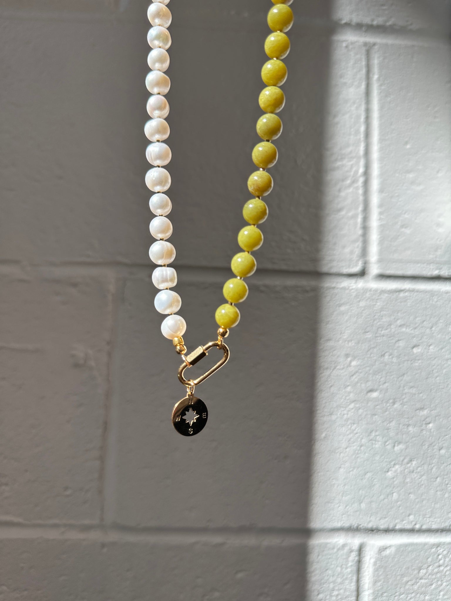 Two toned necklaces on a gray tiled wall, one green and one pearl with a carabiner clasp.